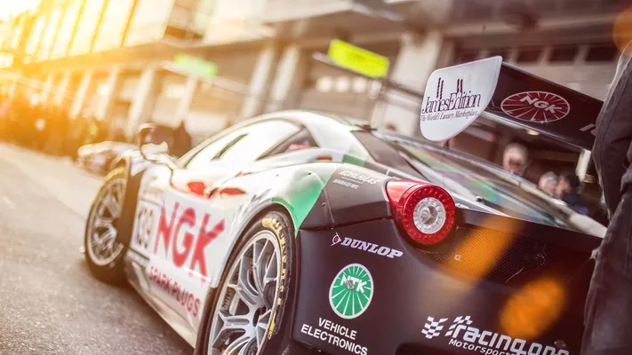 Ein Rennwagen mit großem NGK SPARK PLUGS Logo steht in der sonnigen Boxengasse einer Rennstrecke.