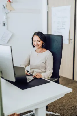Eine lächelnde Mitarbeiterin sitzt an ihrem Schreibtisch in einem modernen Büro und blickt auf ihren Laptop.