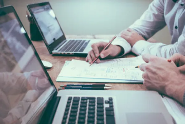 Zwei Personen arbeiten an einem Holztisch, umgeben von Laptops und Dokumenten.
Eine Person hält einen roten Stift und macht Notizen auf einem Blatt Papier, während eine weitere Hand sichtbar ist, die auf ein Dokument zeigt, was eine kollaborative Besprechung oder Arbeitsphase suggeriert.
