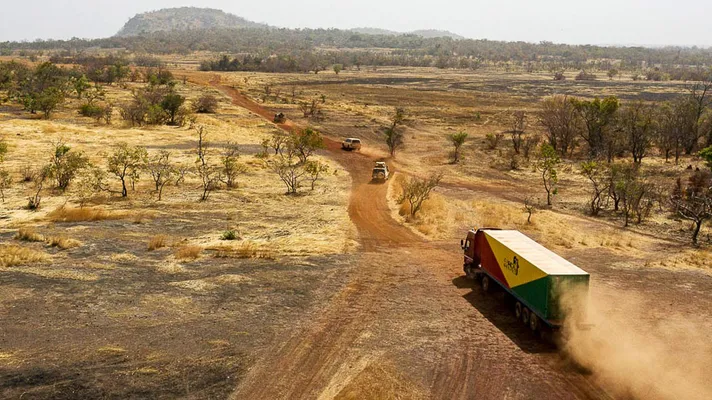 Ein Lastwagen mit einem Solartainer von Africa GreenTec führt einen Konvoi auf einer roten Erdstraße durch eine weite, trockene Savannenlandschaft.