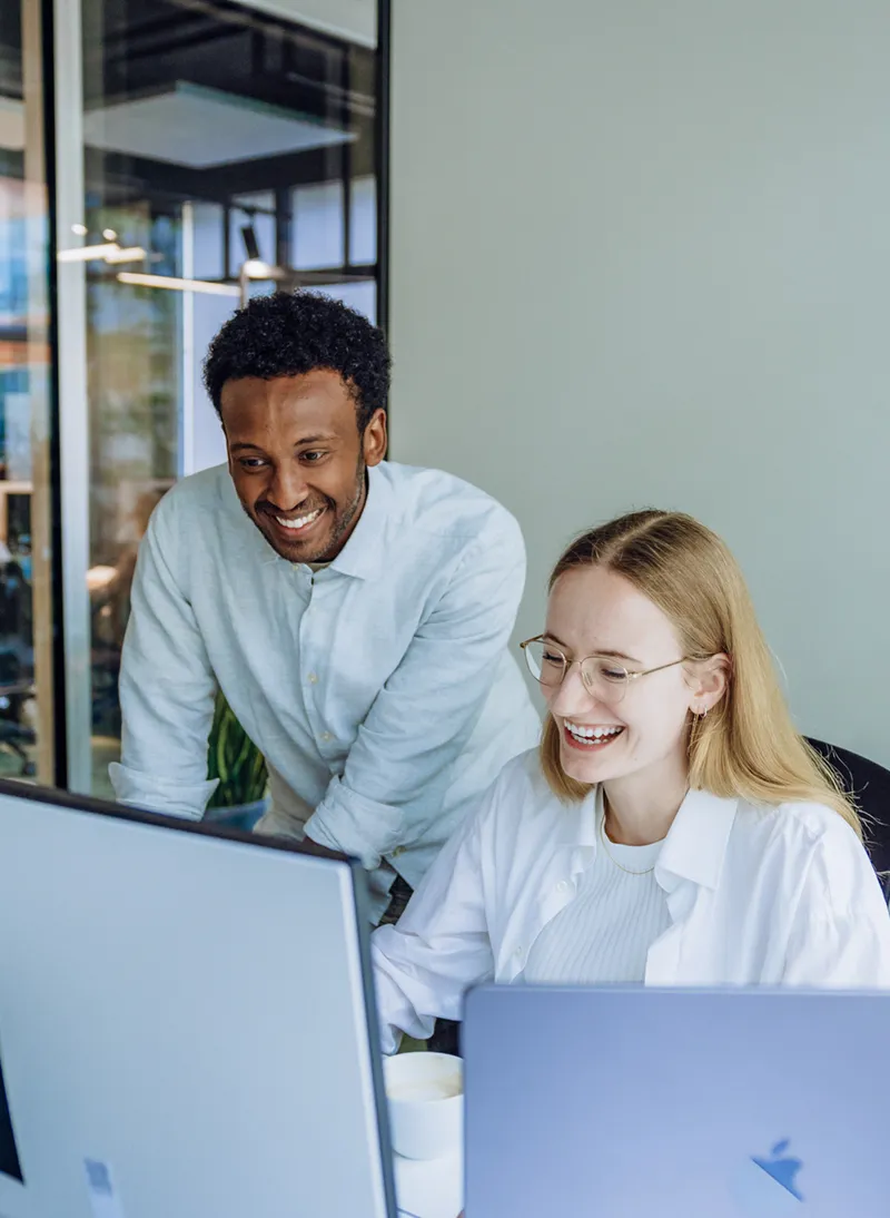Two colleagues smiling and working together at a desk with computers in a modern office setting.