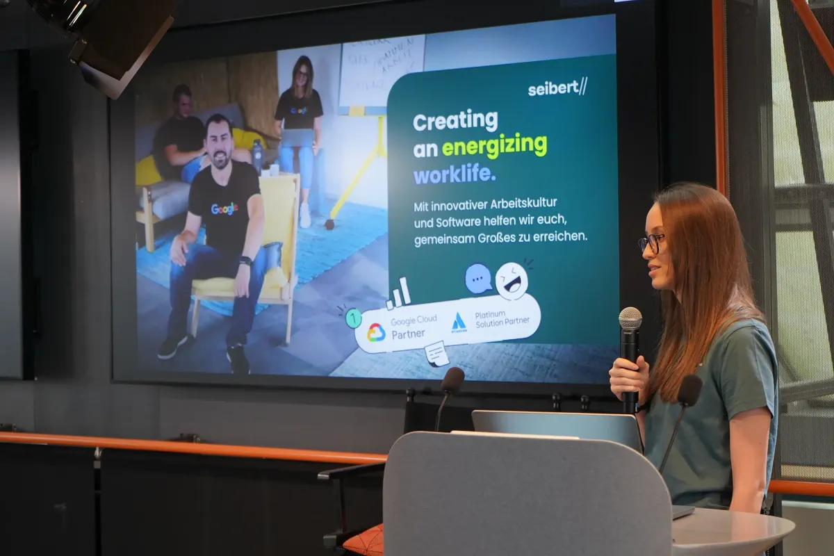Das Foto zeigt eine Emily, die an einem Rednerpult steht und einen Vortrag hält. Sie spricht in ein Mikrofon und blickt aufmerksam ins Publikum. Im Hintergrund ist auf einer großen Leinwand ihre Präsentationsfolie zu sehen. Die Folie zeigt das seibert-Logo, den Slogan Creating an energizing worklife sowie die Logos von Google Cloud Partner und Atlassian Platinum Solution Partner.