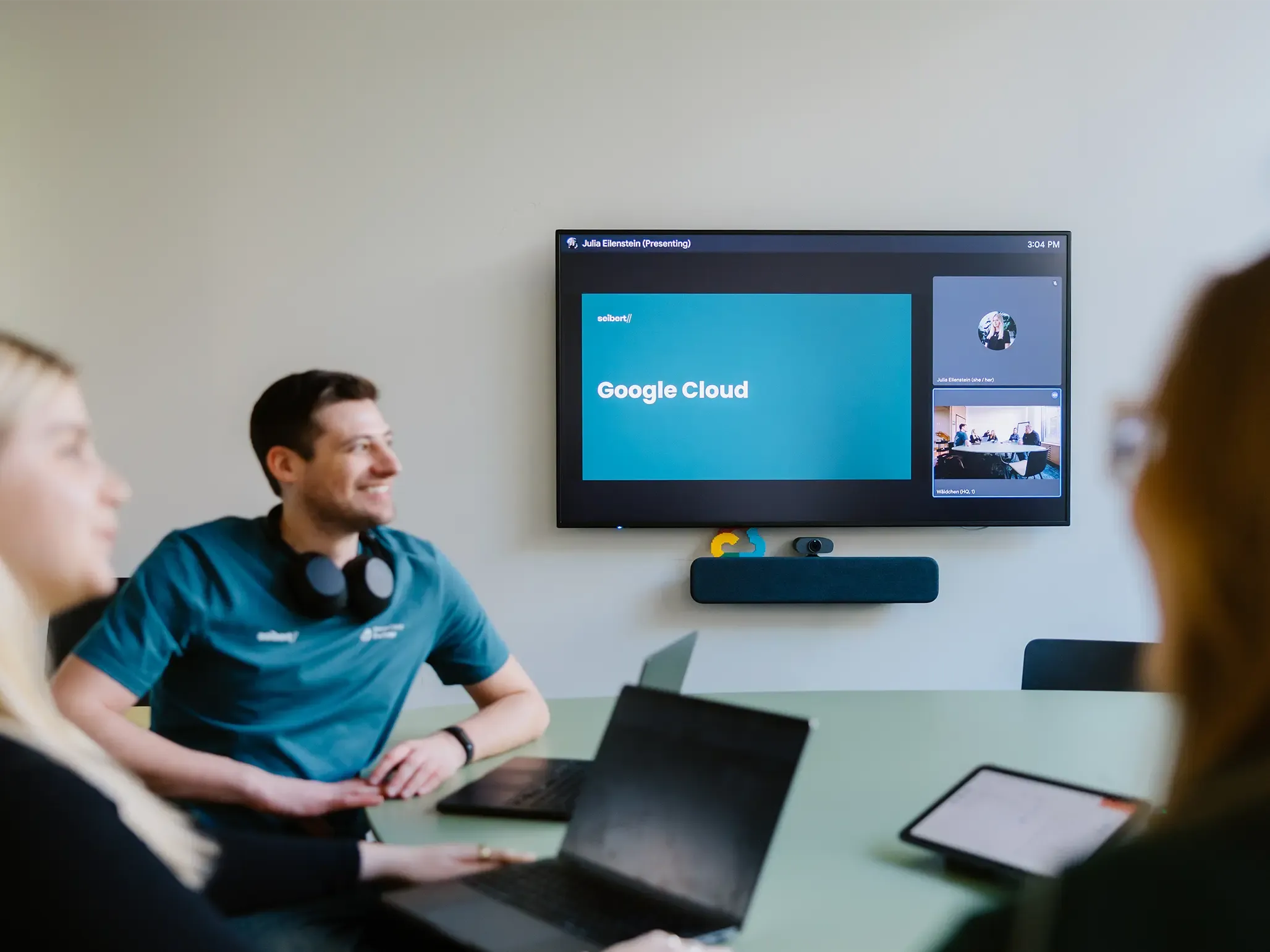 Personen in einem Meetingraum nehmen an einer Videokonferenz teil, bei der auf dem Hauptbildschirm eine Präsentation mit dem Titel Google Cloud gezeigt wird.