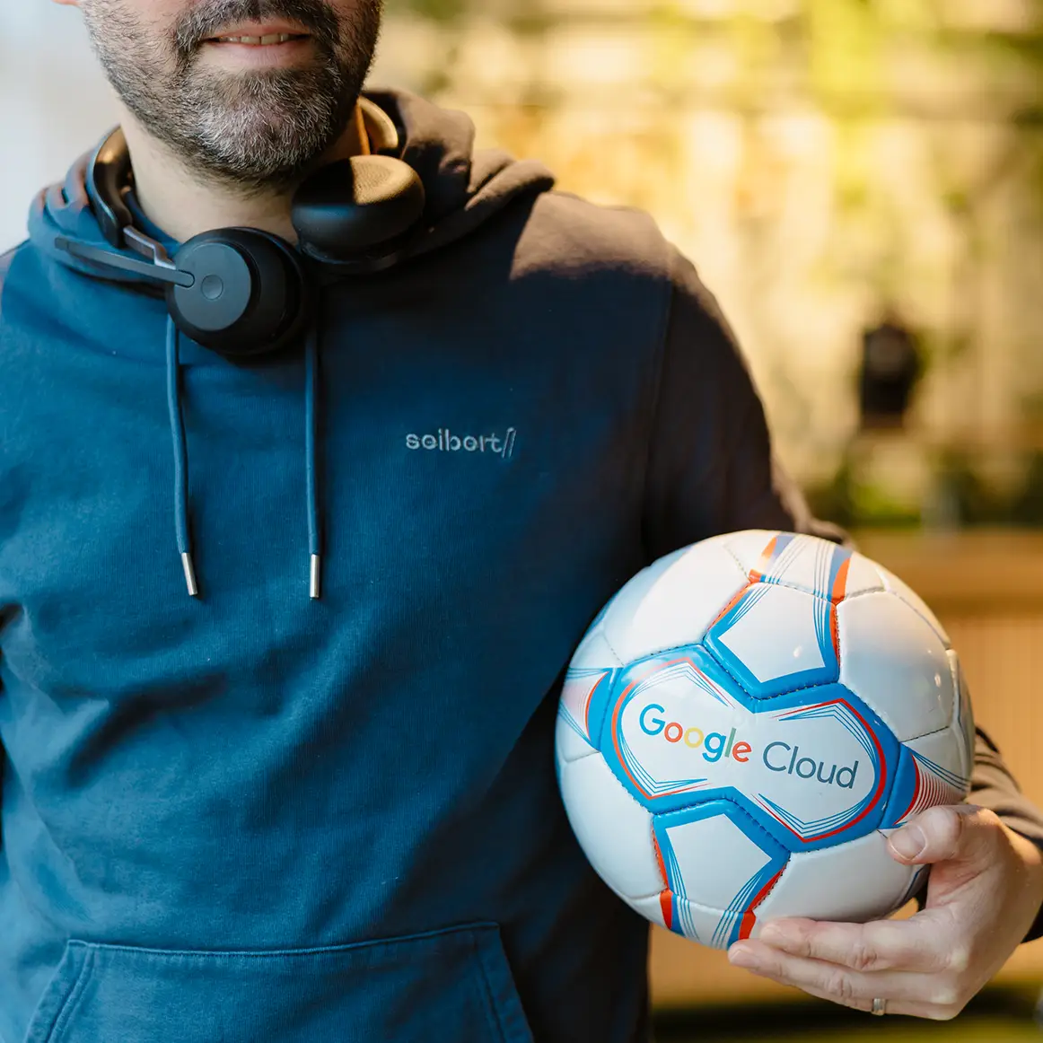 Ein Mitarbeiter im blauen Hoodie mit Seibert-Logo hält einen Fußball mit dem Google Cloud Logo.