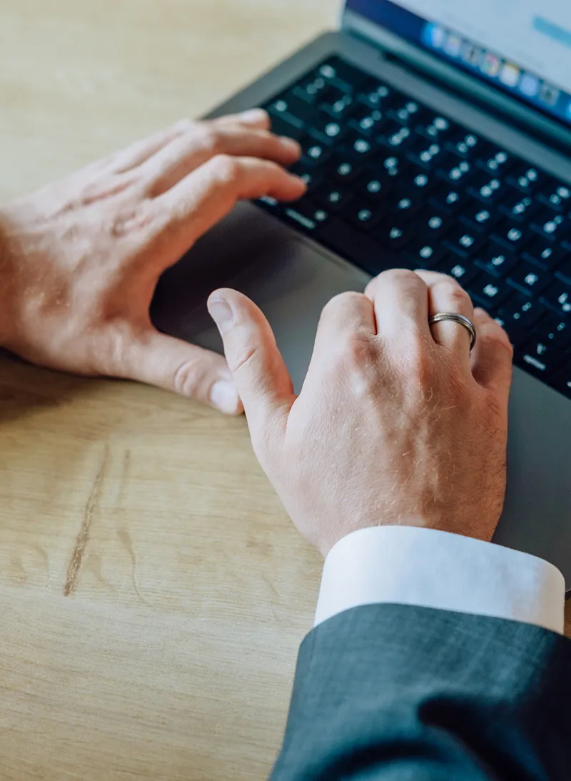 Close-up of hands typing on a laptop keyboard, wearing a suit and ring.