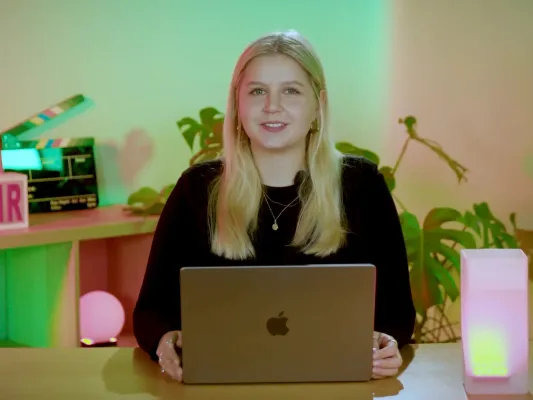 Eine Expertin sitzt lächelnd mit einem Laptop am Schreibtisch in einer modernen Umgebung für einen Online-Kurs.