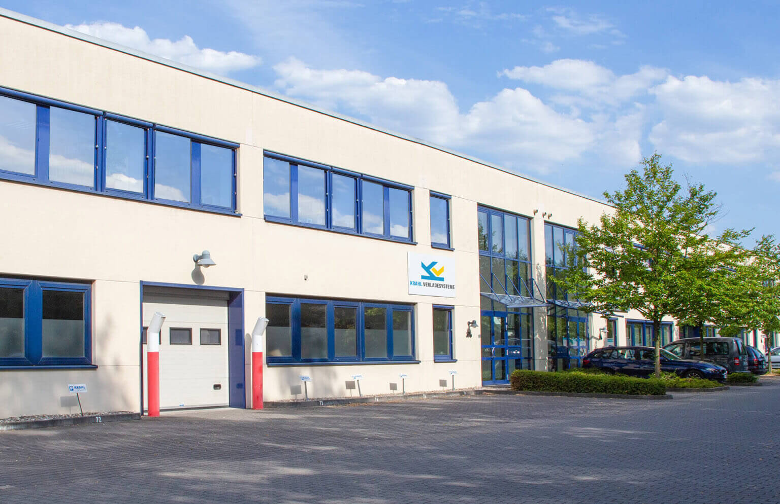 Die Außenansicht des Firmengebäudes von Krahl Verladesysteme an einem sonnigen Tag. Das helle, moderne Gebäude hat blaue Fensterrahmen und davor befinden sich Parkplätze mit Bäumen.