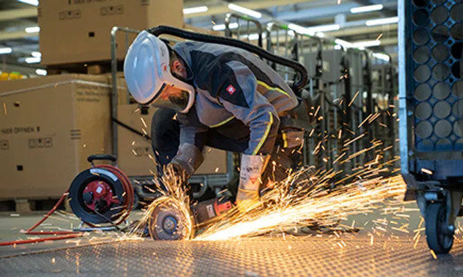 Ein Arbeiter in voller Schutzausrüstung mit Helm und Atemschutz kniet auf einem Gitterrost und bearbeitet Metall mit einem Winkelschleifer, wobei ein heller Funkenregen entsteht.