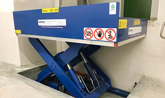 Ein blauer Scherenhubtisch der Marke Krahl, der zur Hälfte in eine Grube im Betonboden abgesenkt ist. Auf der Plattform sind Warnhinweise und das Firmenlogo sichtbar.