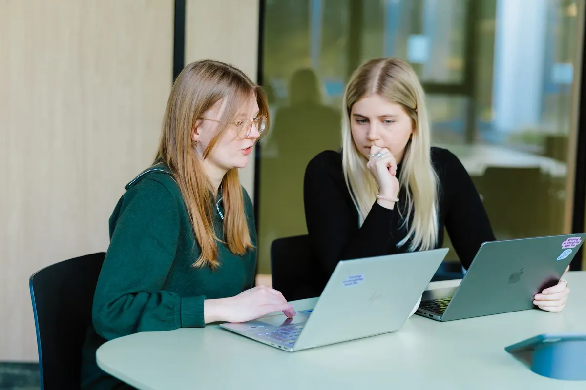 Das Foto zeigt zwei Lara und Julia, die nebeneinander an einem Tisch sitzen und jeweils an einem Laptop arbeiten. Die Lara gestikuliert in Richtung ihres Bildschirms erklärt etwas. Julia hört ihr nachdenklich und konzentriert zu. Die Szene vermittelt eine Atmosphäre von professioneller Zusammenarbeit, Wissensaustausch oder Mentoring.