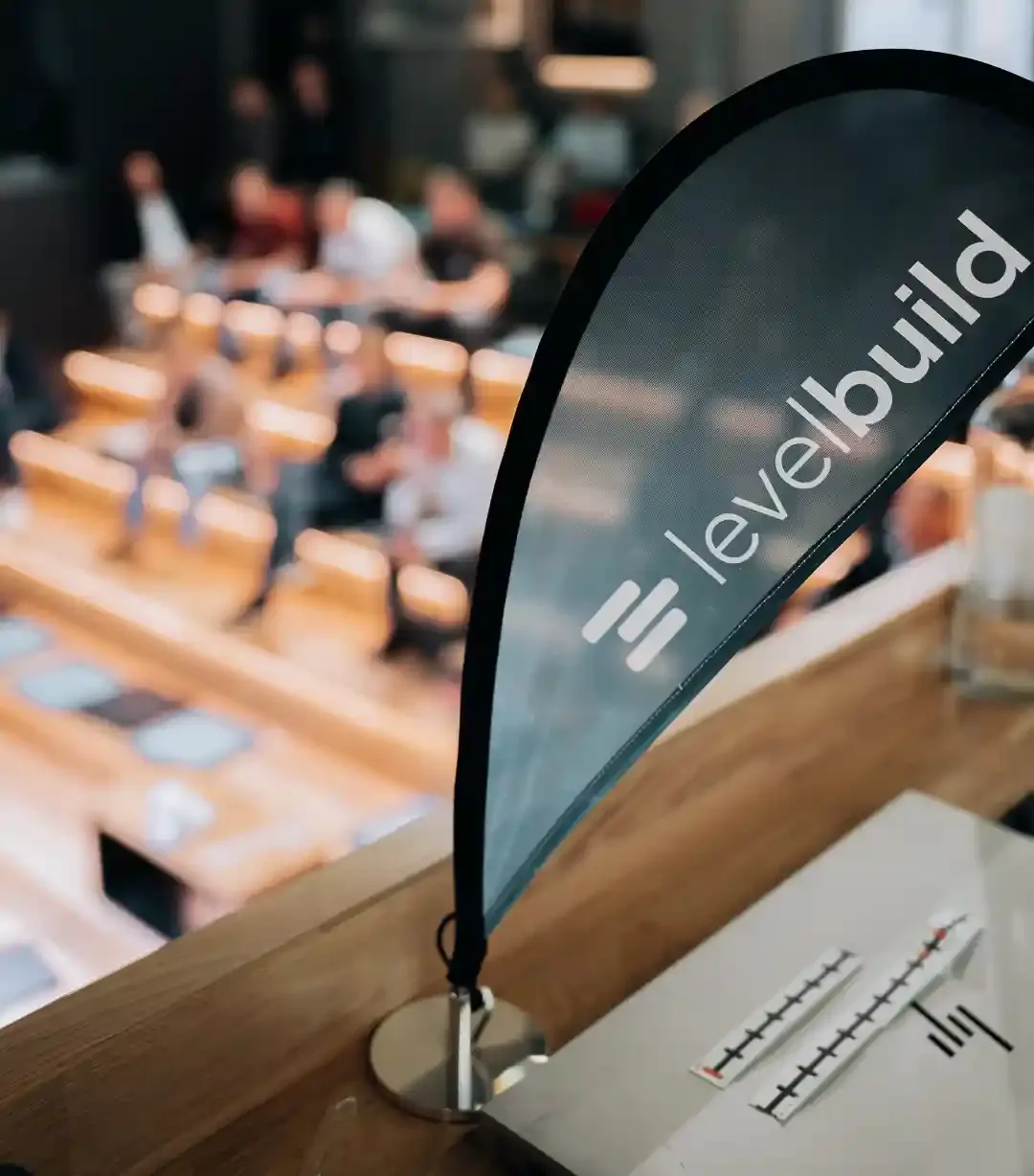 Eine kleine schwarze Levelbuild-Fahne steht im Vordergrund auf einem Holztresen. Im unscharfen Hintergrund ist ein Publikum bei einer Konferenz oder einem Vortrag zu sehen.