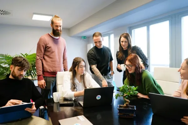 Eine Gruppe von jungen Mitarbeitern in einem hellen Besprechungsraum, die sich um einen Laptop versammelt haben und gemeinsam an einem Projekt arbeiten. Die Atmosphäre wirkt kollaborativ und kreativ.