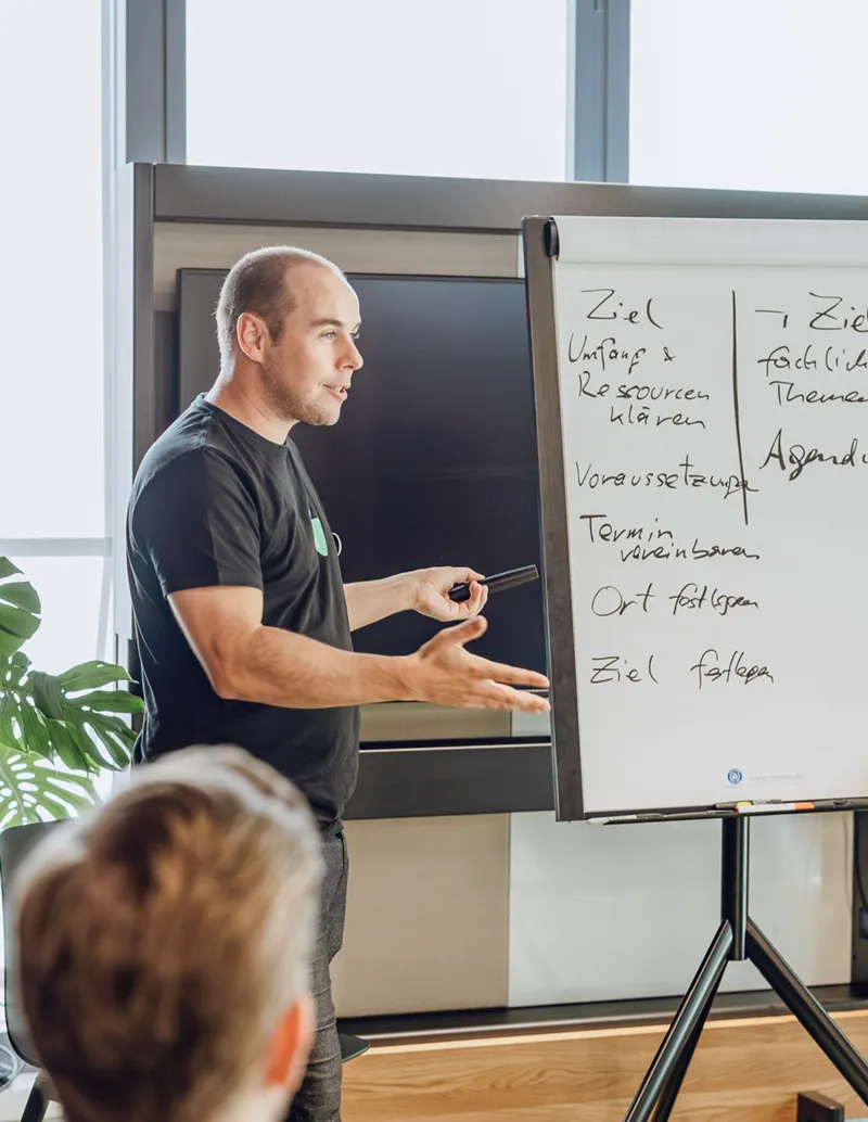 A man presenting in an office setting, standing next to a flipchart with handwritten notes. Several people are seated, listening attentively. Large windows and plants are visible in the background.