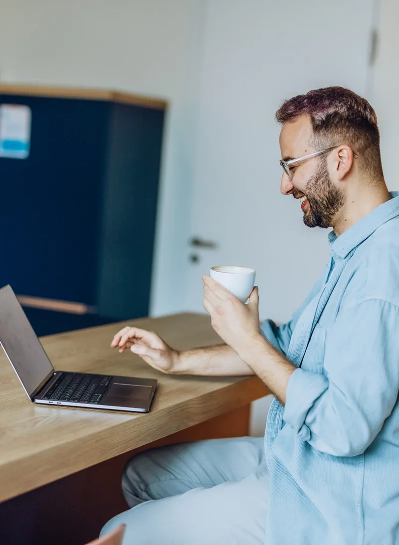Man sitting at a kitchen counter with a laptop and holding a coffee cup, smiling while looking at the screen.