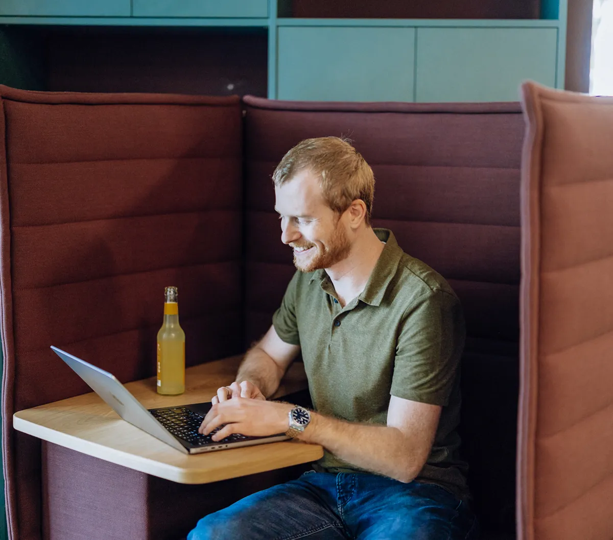 A man sitting in a cozy booth working on a laptop with a bottle of drink on the table.