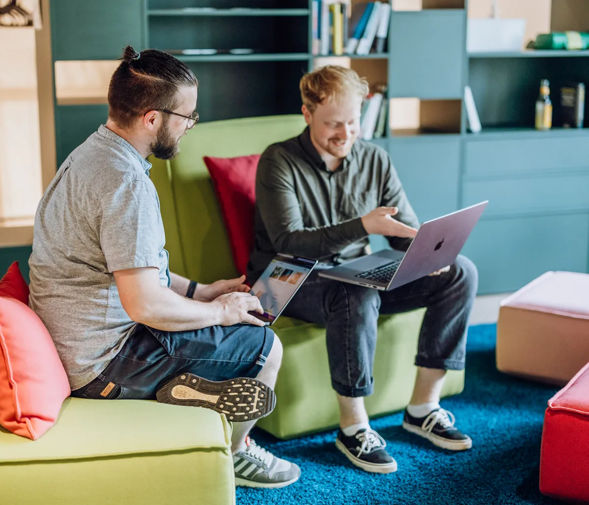 Two men sitting on colorful couches using laptops in a modern office setting.