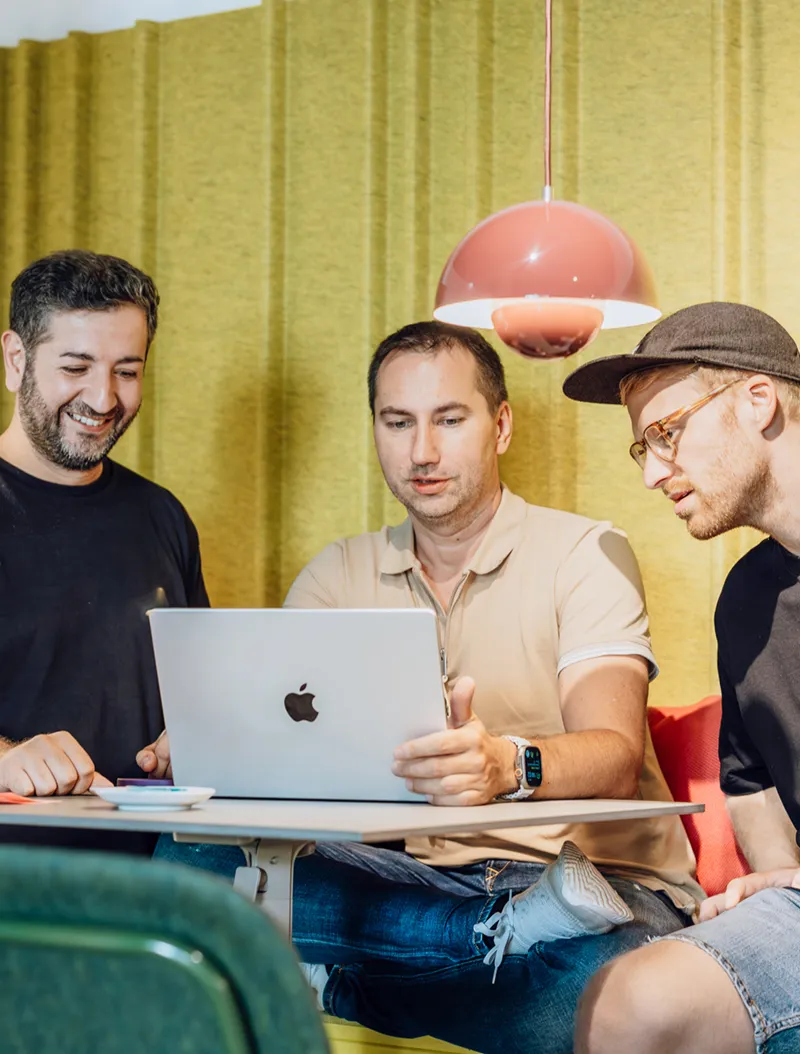 Three men sitting at a table, looking at a laptop screen, with a yellow background and red pendant lights.
