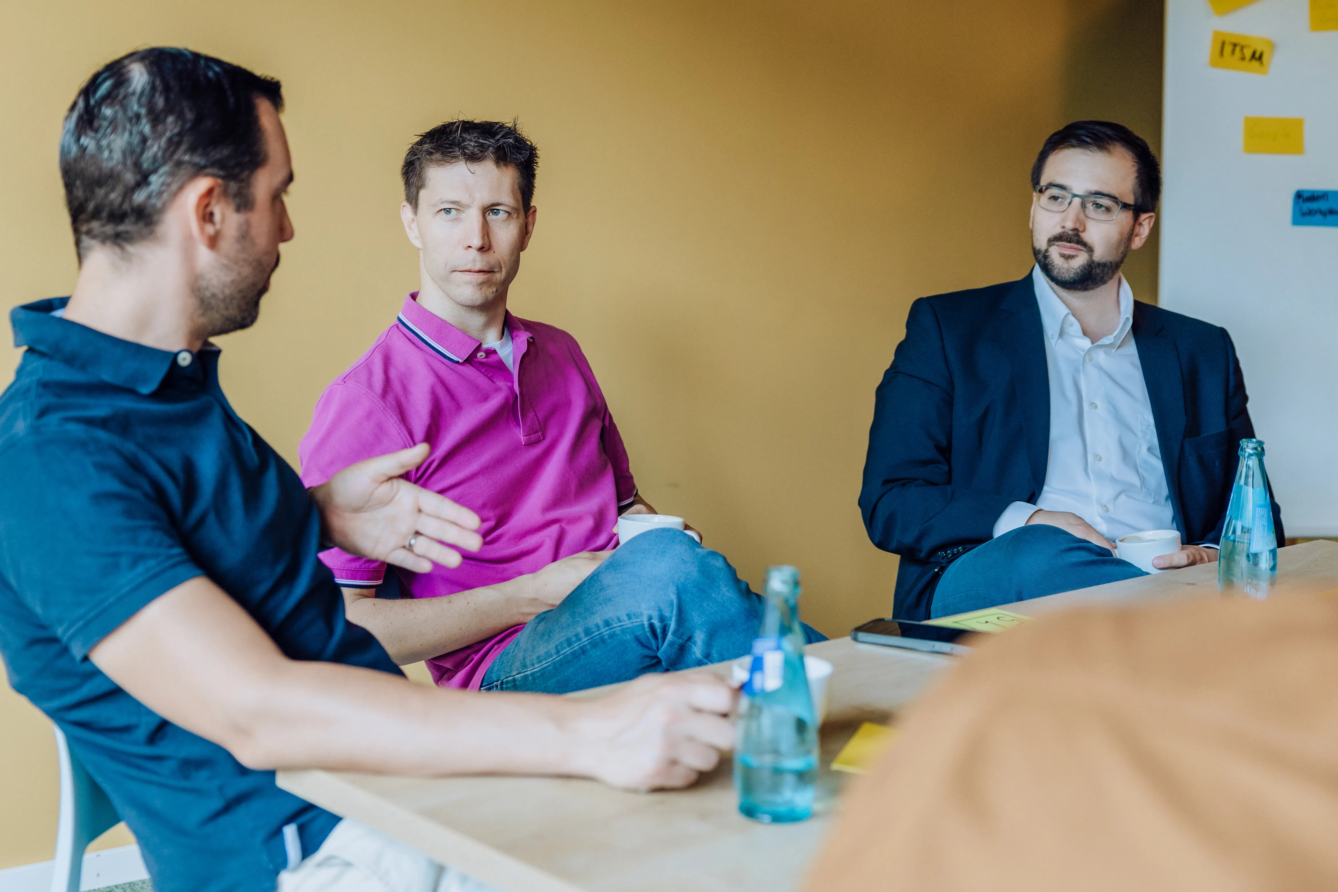 Three men sitting at a table having a discussion with water bottles and sticky notes visible.