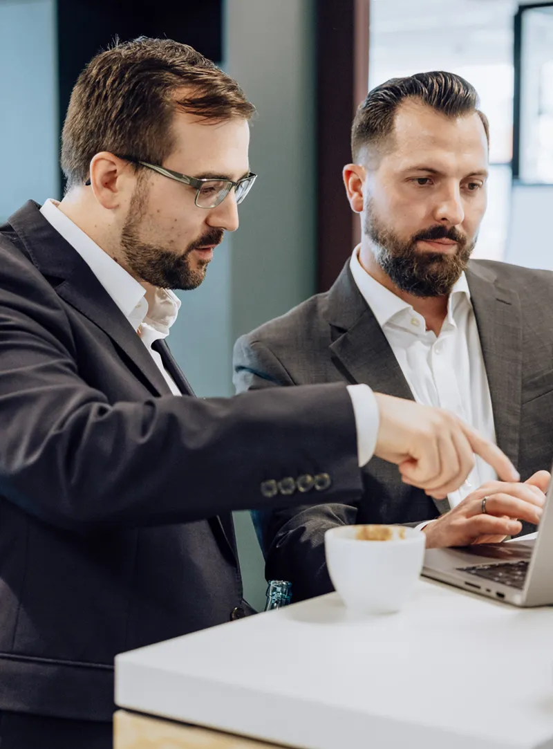 Two men in suits discussing something on a laptop at a table with a coffee cup.