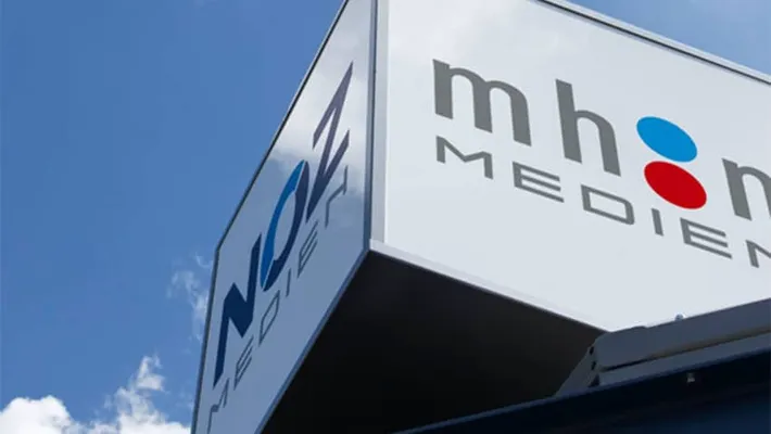 Das weiße Firmenschild mit den Logos von NOZ MEDIEN und mhn MEDIEN an einem Gebäude, aufgenommen aus einer Froschperspektive gegen den blauen Himmel.