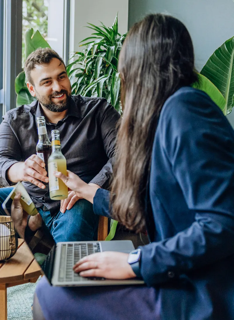 Two people sitting in an office setting with plants, clinking bottles and smiling, one using a laptop.