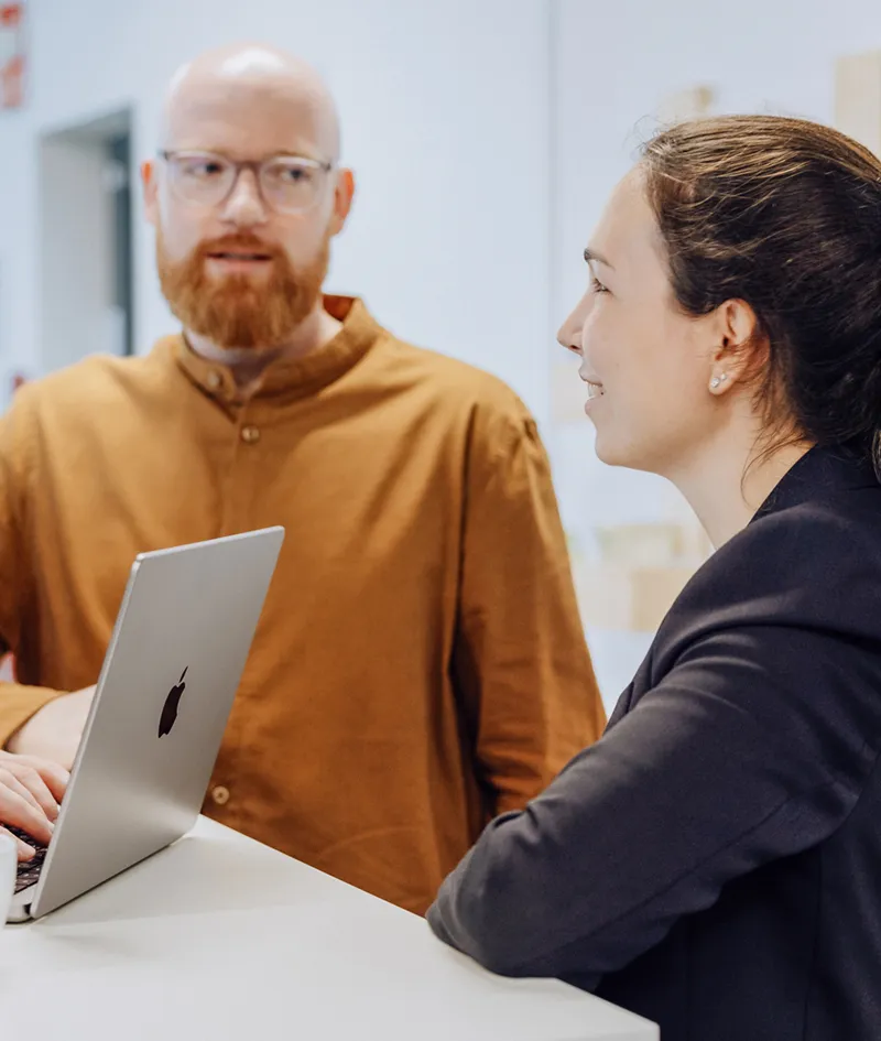 Two people having a conversation at a high table with a laptop and a cup of coffee, in a modern office setting.