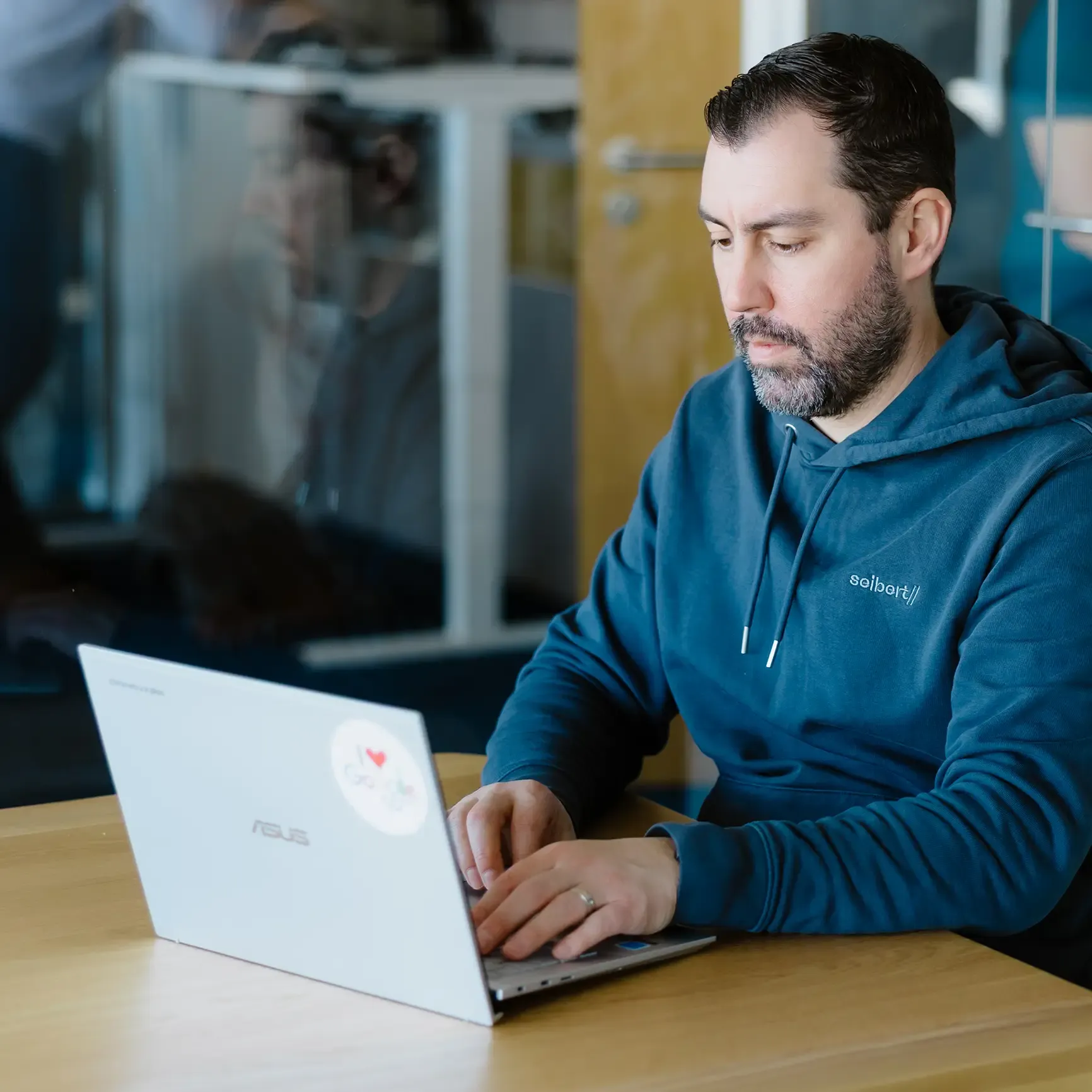 Ein Mitarbeiter im blauen Seibert-Hoodie arbeitet konzentriert an einem silbernen Laptop, auf dem ein "I love Google"-Sticker angebracht ist.