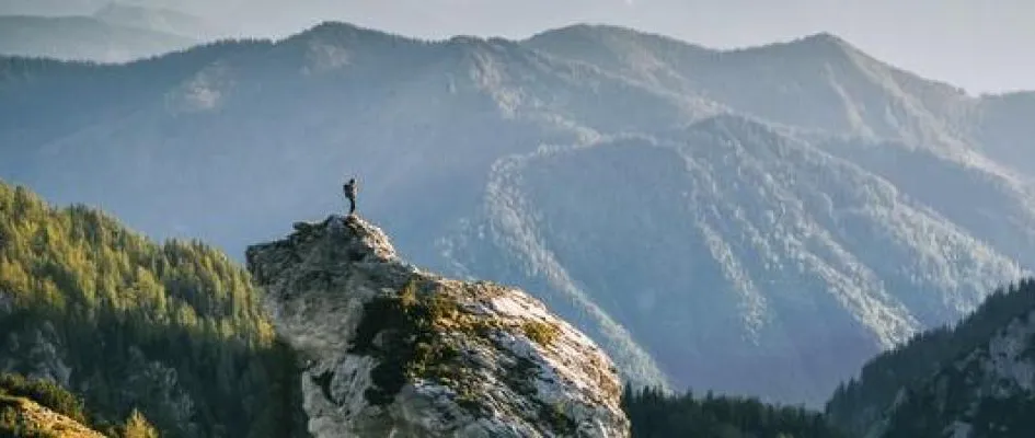 Ein Wanderer steht allein auf einem Felsgipfel und blickt über eine weite, neblige Berglandschaft, was ein Gefühl von Weitsicht und Erfolg vermittelt.