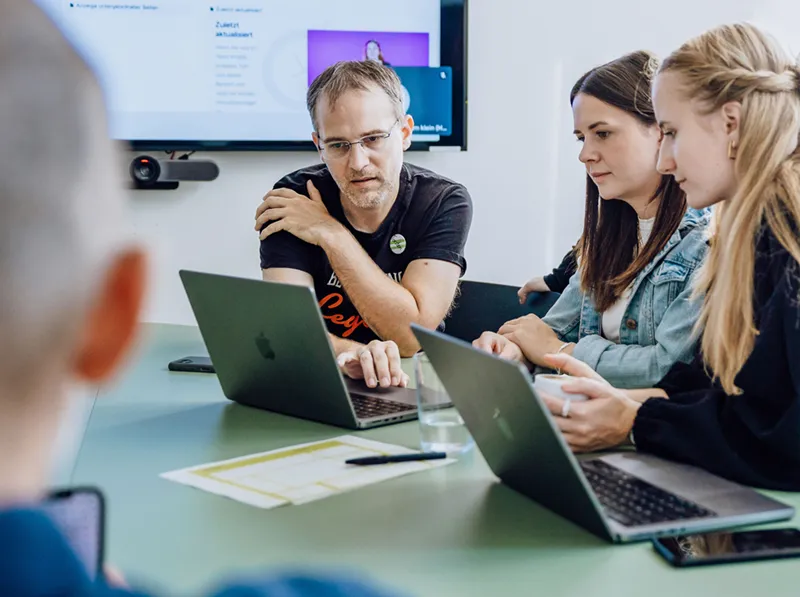 A group of people sitting at a table with laptops, engaged in a discussion. A screen is visible in the background displaying a presentation.