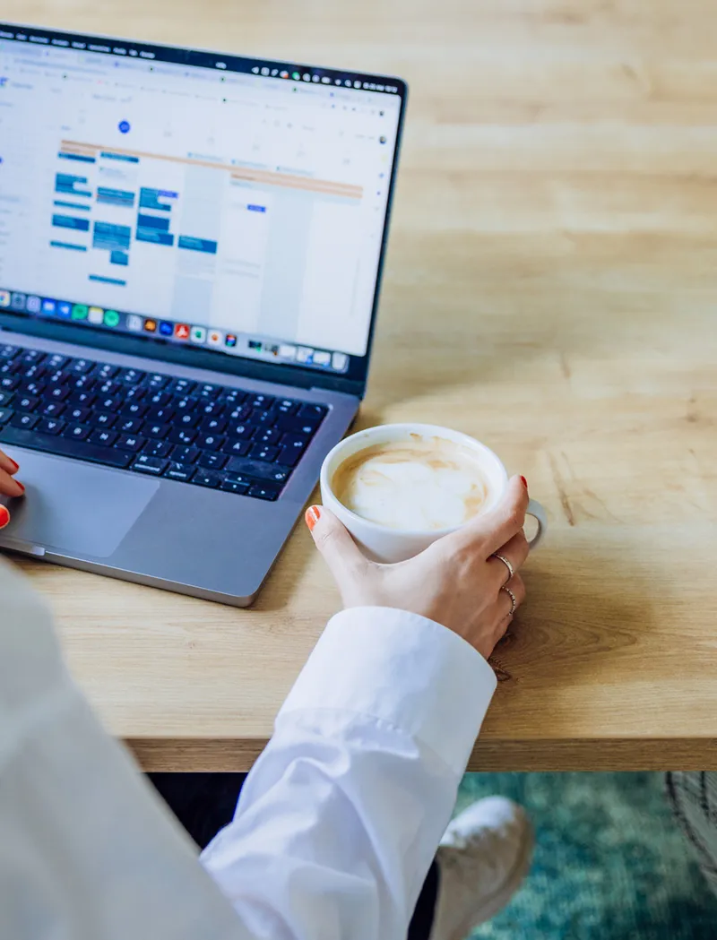Two people working on laptops at a wooden table, one holding a coffee cup, with a video call on one screen.