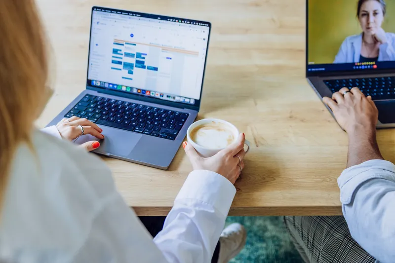 Two people working on laptops at a wooden table, one holding a coffee cup, with a video call on one screen.