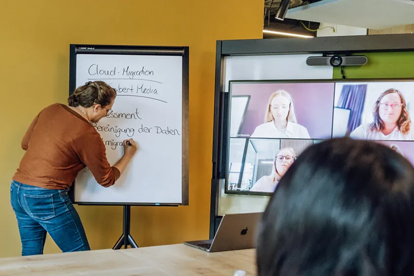 A woman writing on a flipchart during a video conference with four participants displayed on a screen.