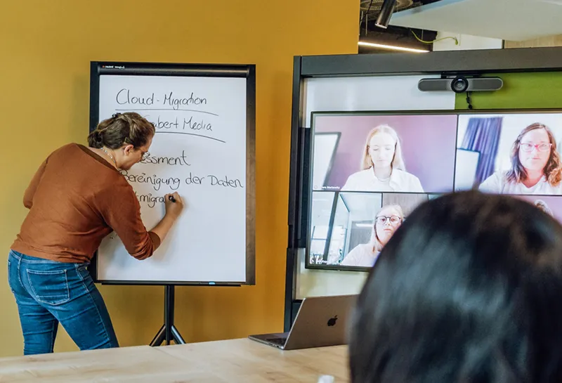 A woman writing on a flipchart during a video conference with four participants displayed on a screen.
