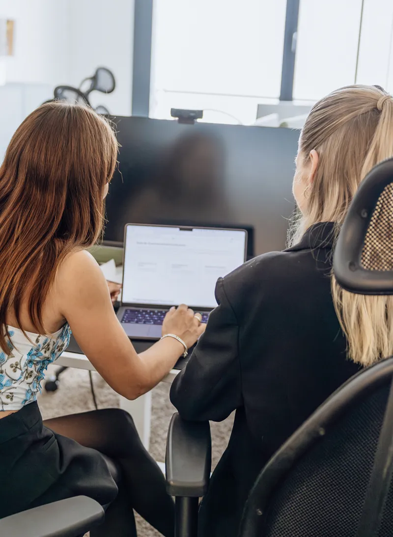 Two women collaborating at a desk, looking at a laptop screen in an office setting.