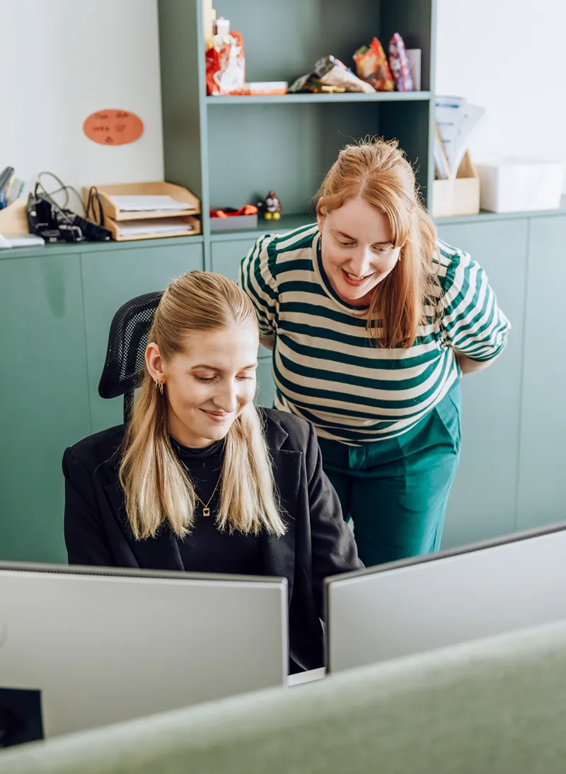 Two women in an office setting, one sitting at a desk with a computer and the other standing beside her, both smiling and looking at the screen.